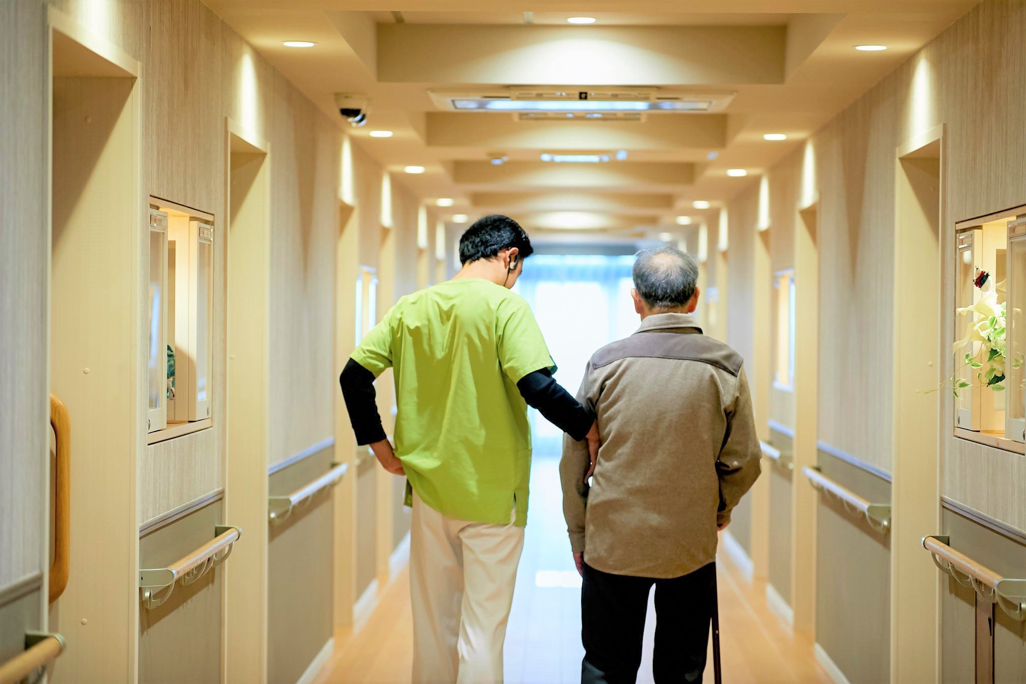 介護スタッフが入居者の歩行をサポートする廊下。清潔で明るい共用部の実風景。